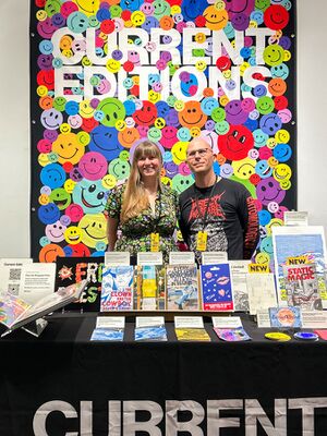 Two people are standing behind a table covered with publications at a vibrant event. The woman on the left is wearing a floral dress, and the man on the right is in a black shirt with red text that reads “INSECT WARFARE”. Behind them is a colorful, eye-catching backdrop filled with smiley faces in various colors, with the words "CURRENT EDITIONS" prominently displayed at the top.